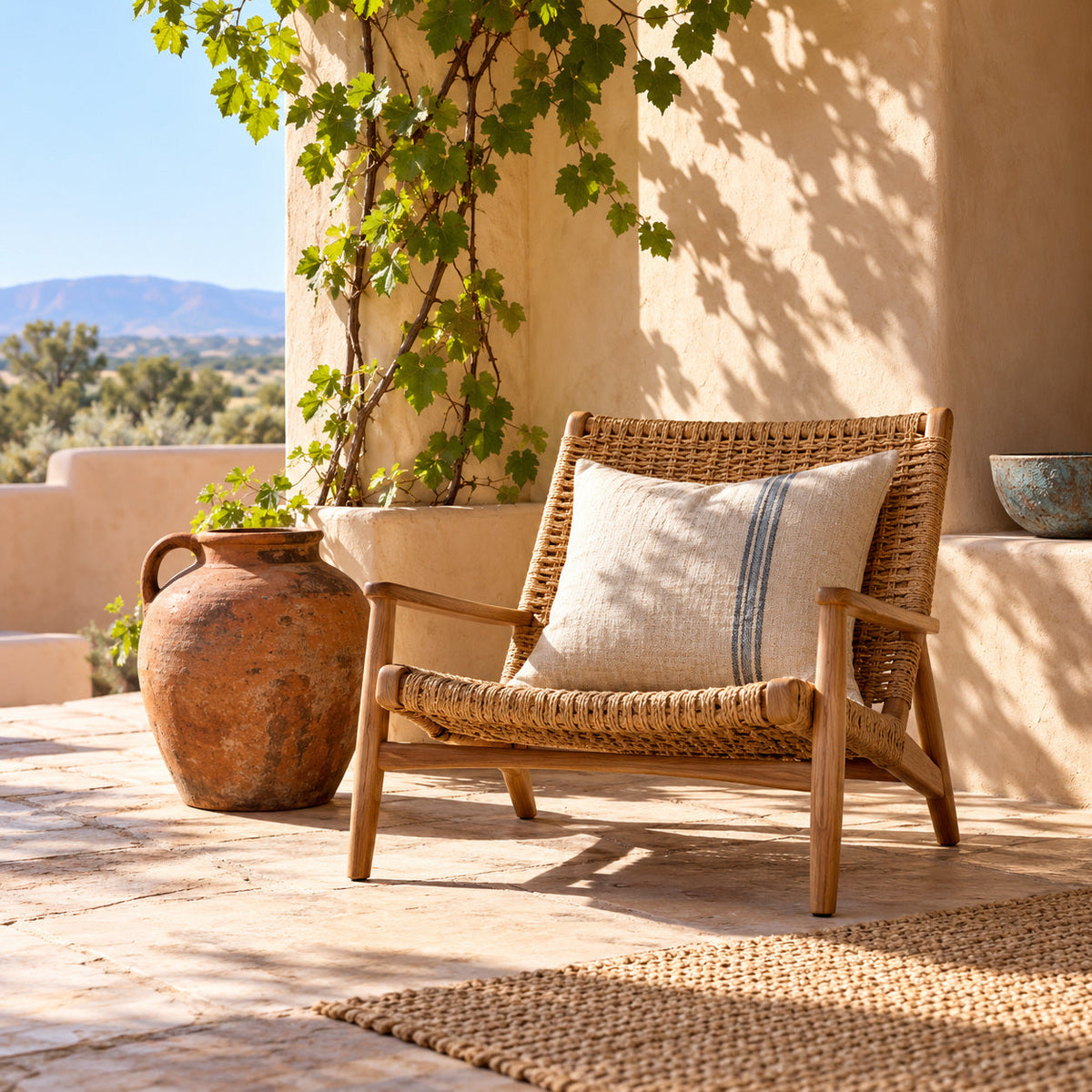 Woven chair, clay water jug and linen cushion in a sunlit adobe portal with vine shadows.