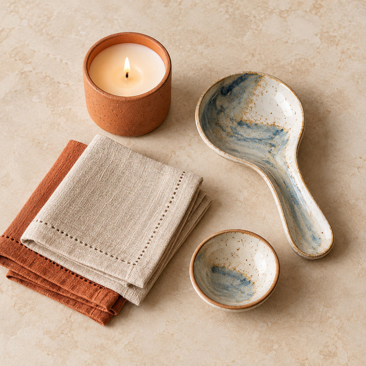 Flat lay of a clay candle, ceramic spoon rest, linen napkins and small ceramic dish.