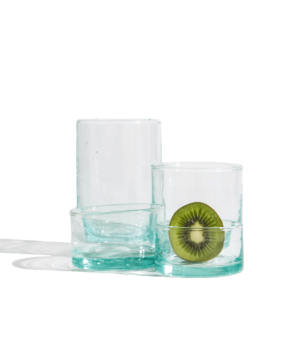 Three sizes of hand-blown recycled glass tumblers stacked with a halved kiwi on a deep green ground