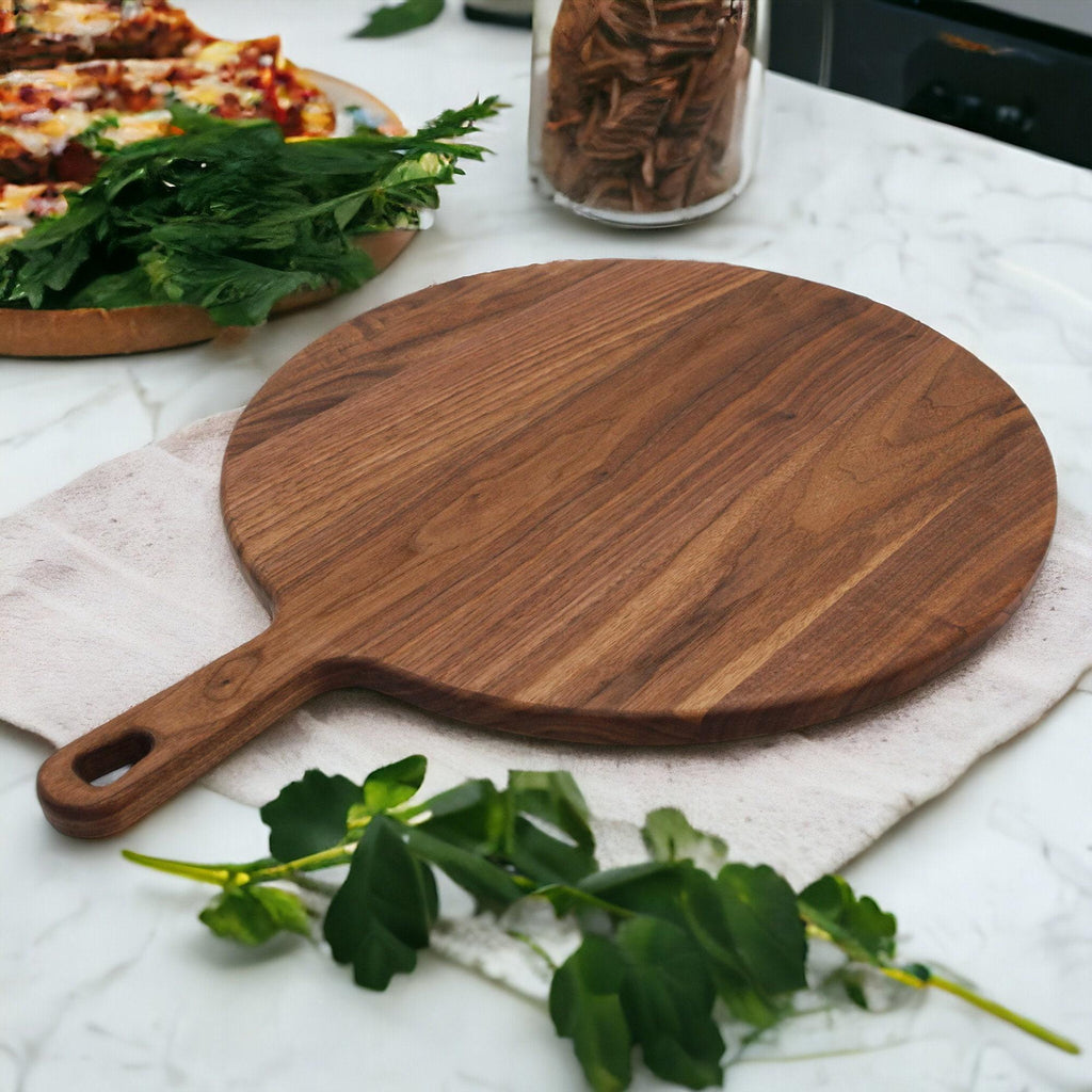 Round walnut pizza paddle on a marble counter with fresh parsley, a pizza and a jar of pasta behind