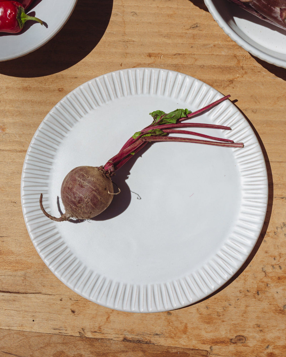 Matte white ceramic dinner plate with a carved rim on a worn wood table, a single beet laid across it