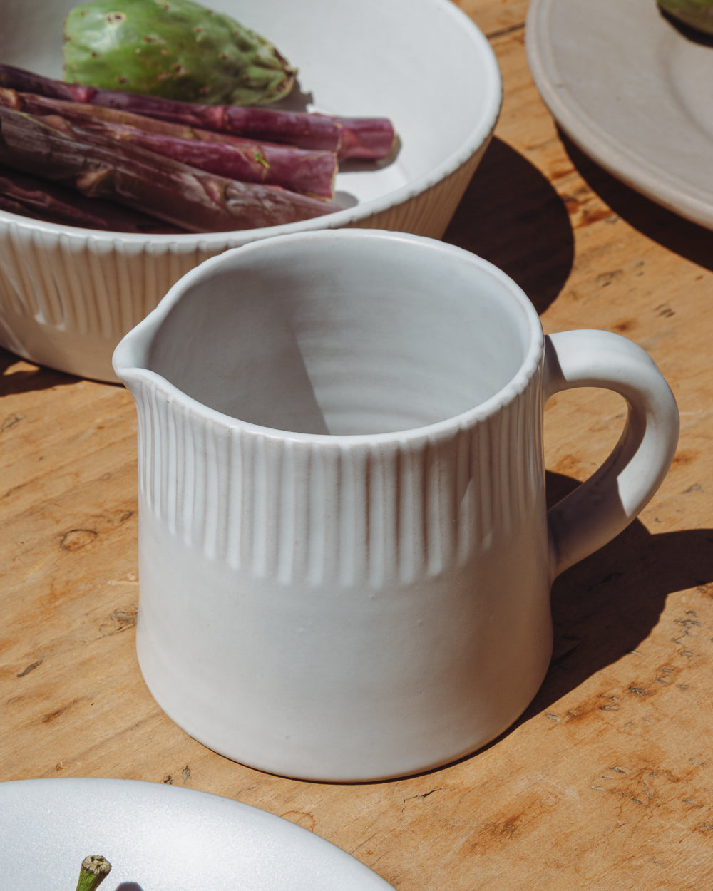 Small carved white ceramic pitcher on a wood table beside a bowl of purple asparagus