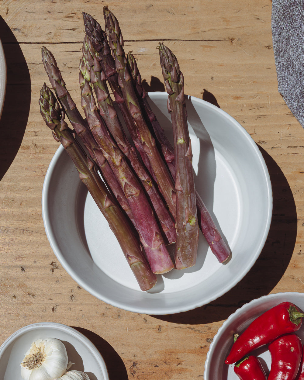Purple asparagus in a matte white carved ceramic bowl next to red chiles on a wood table