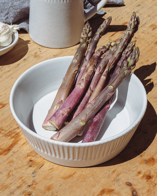 Matte white carved serving bowl holding purple asparagus on a worn wood table, seen from above