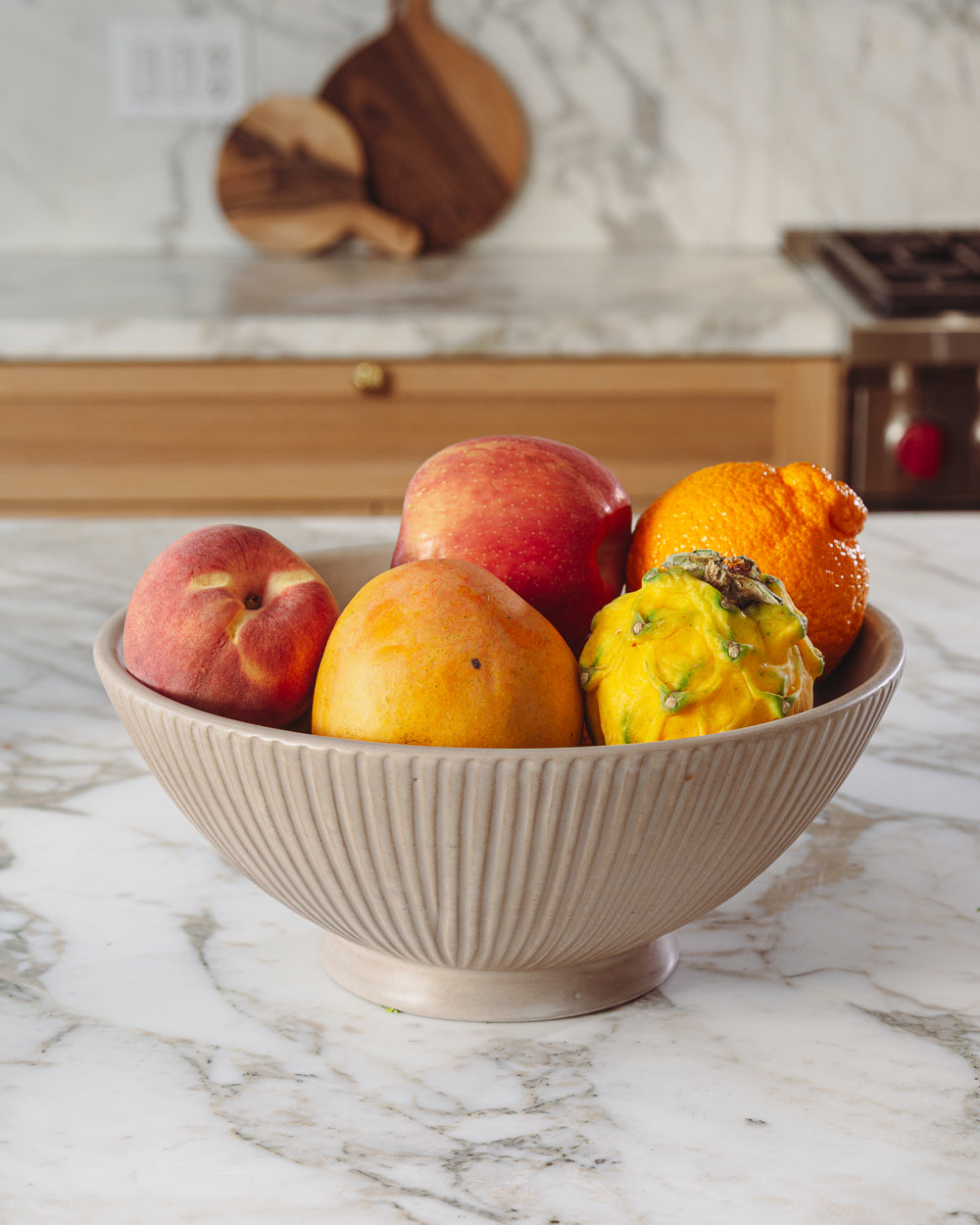 Carved clay-toned serving bowl filled with peaches and small squash on a white marble kitchen counter