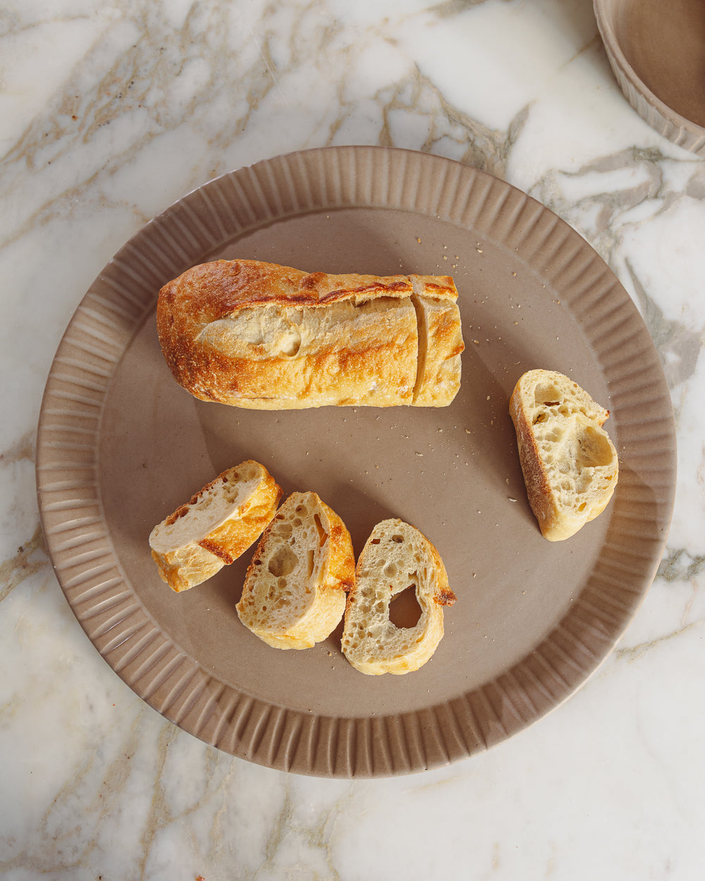 Clay-toned ceramic platter with a carved fluted rim holding a sliced baguette on a white marble counter