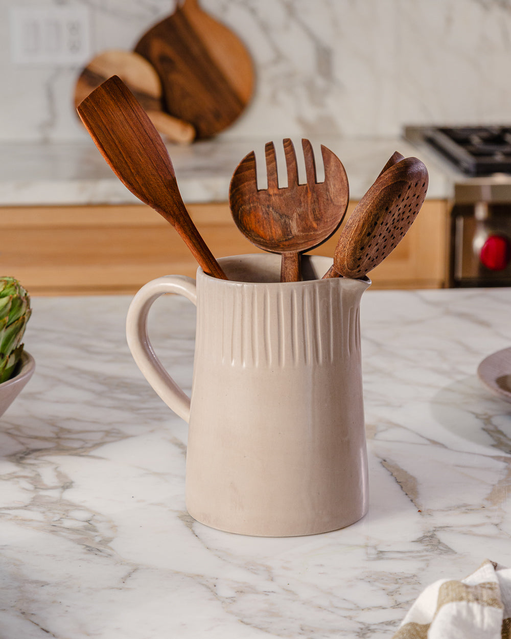 Large carved clay-toned pitcher filled with wooden utensils on a white marble kitchen counter
