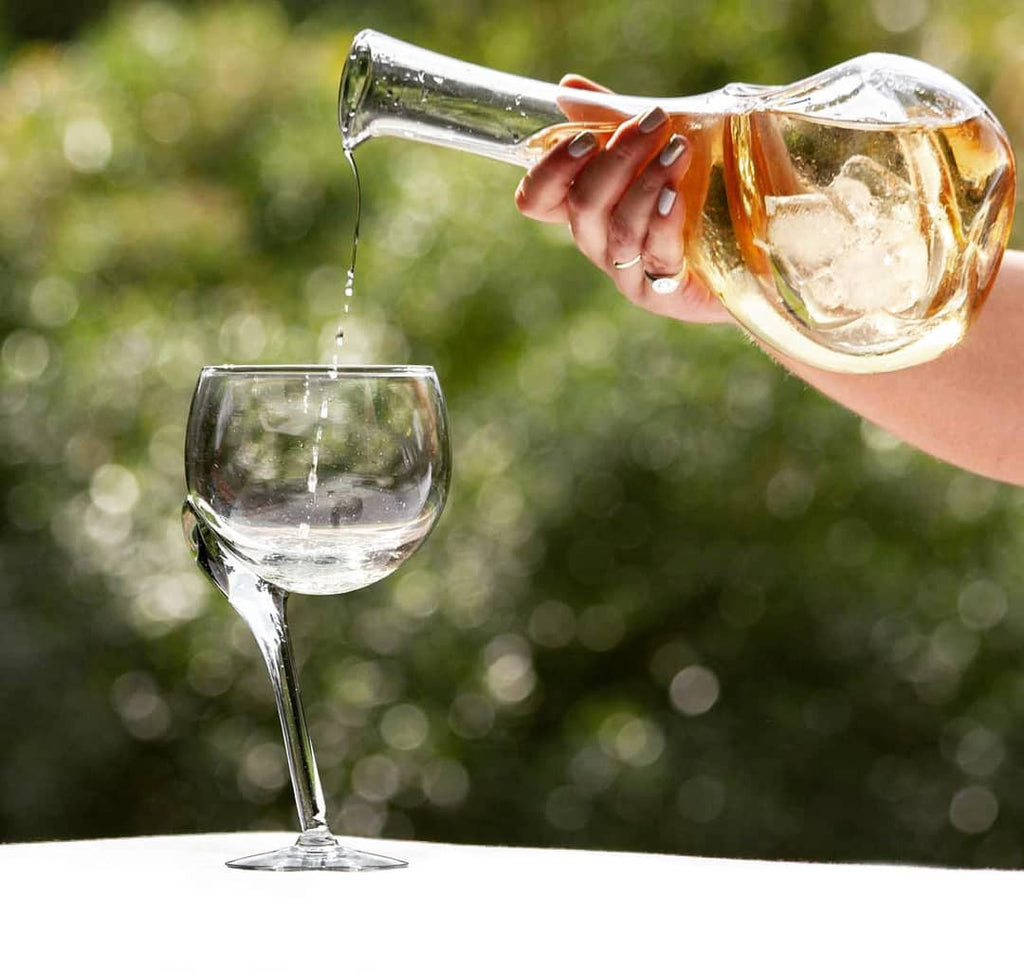 Hand pouring chilled white wine from a recycled glass decanter into a wine glass in a garden