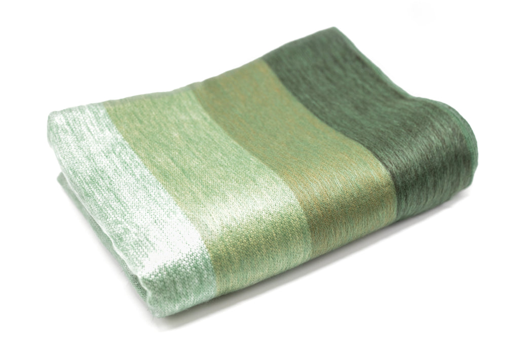 Folded sage and juniper striped alpaca throw showing its pale finished edge on a white background