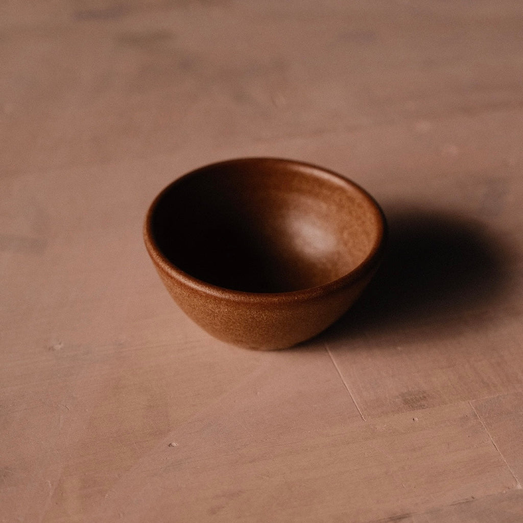 Small stoneware pinch bowl in a warm rust-brown glaze casting a long shadow on a sand-colored surface