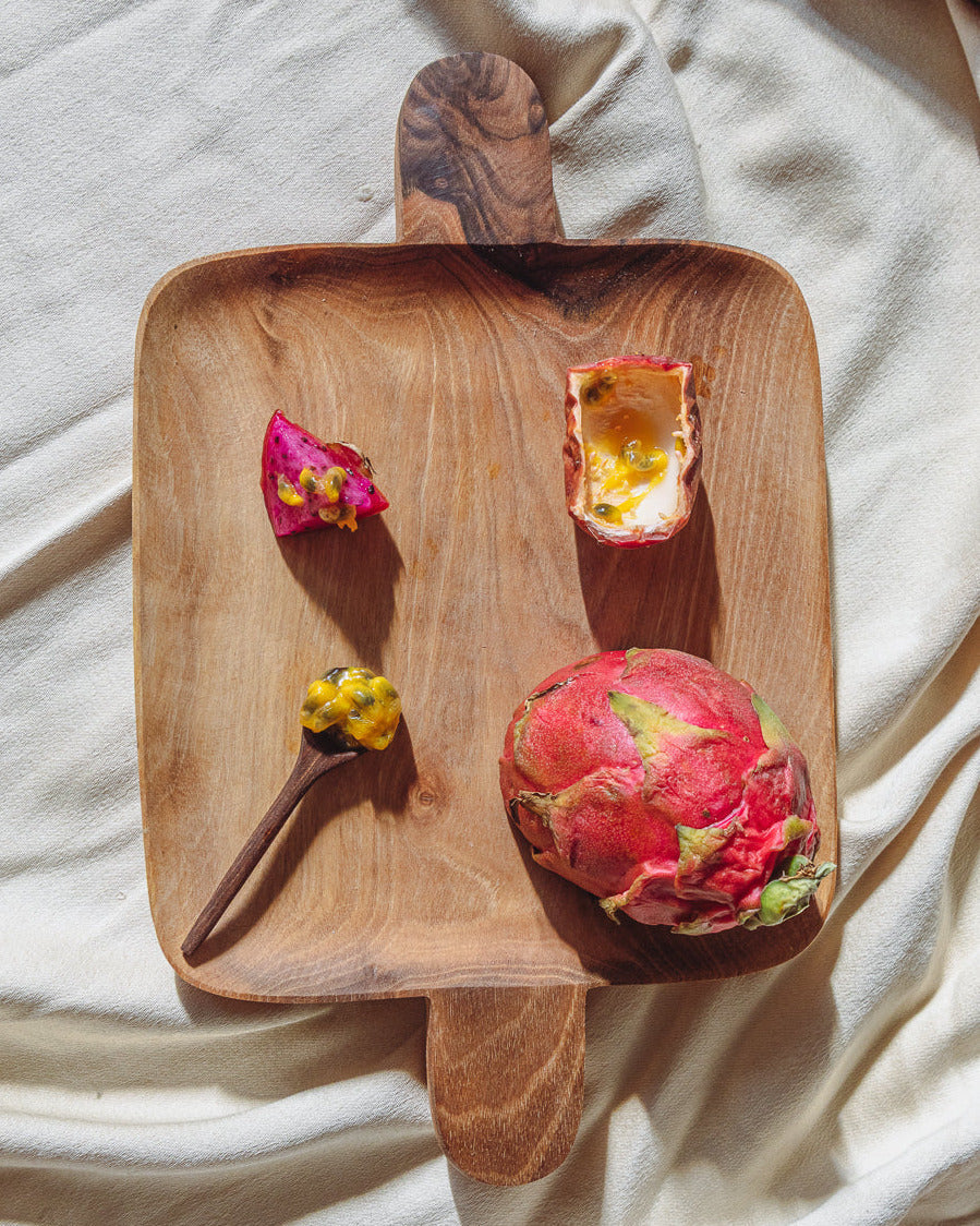 Walnut serving tray on rumpled linen holding a dragon fruit, passion fruit halves and a sprig of herbs