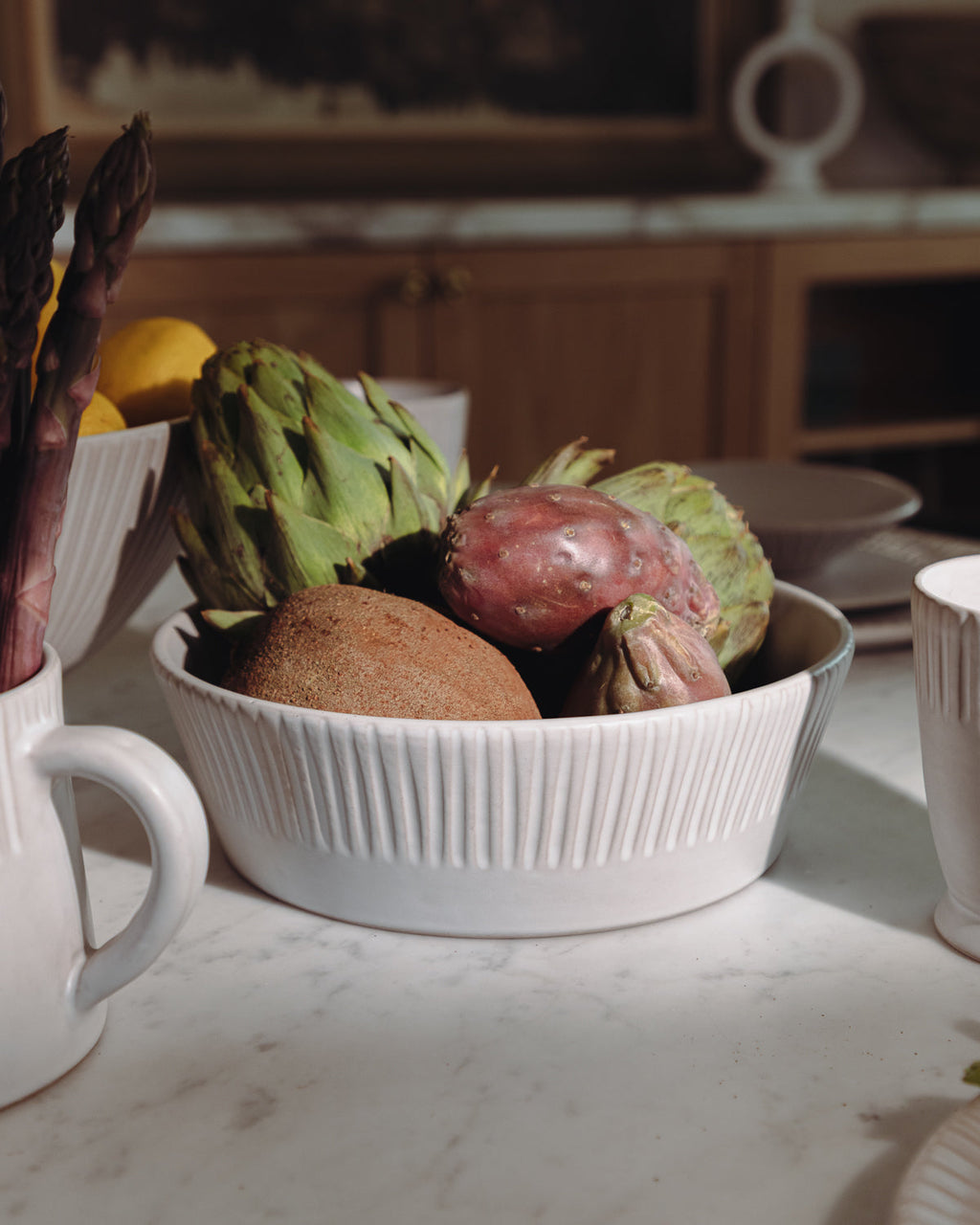 Carved white ceramic serving bowl of artichokes and vegetables on a kitchen counter with a pitcher