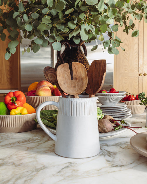 Large white carved pitcher holding wooden spoons on a marble counter beside a bowl of vegetables