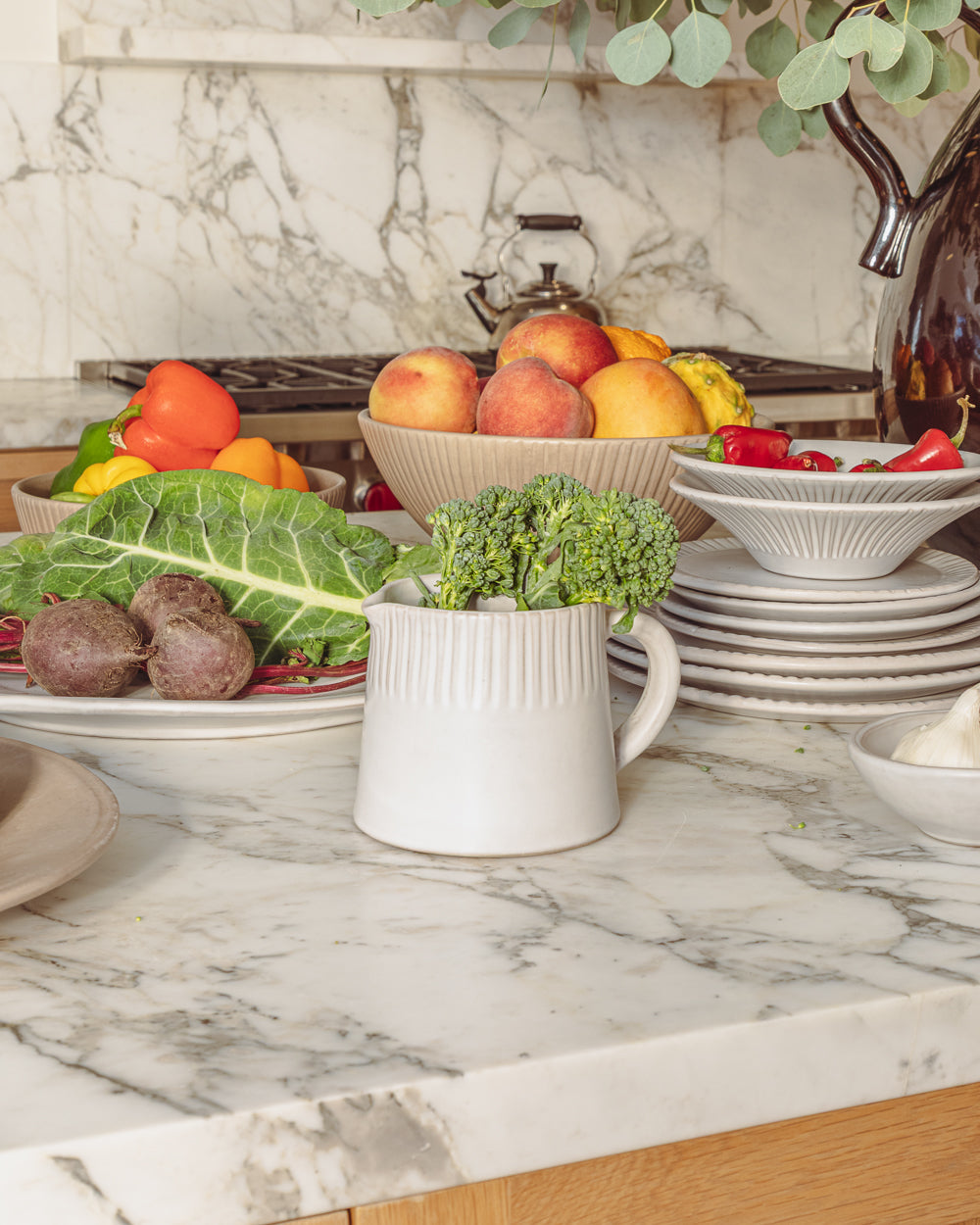 Carved white ceramic pitcher on a marble counter surrounded by fruit, greens and stacked plates