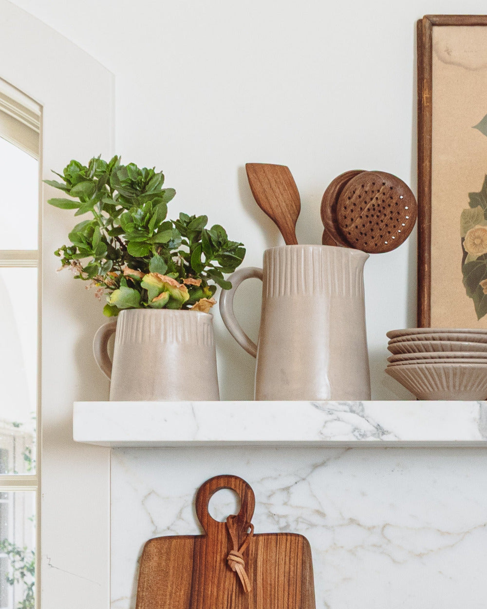 Clay-toned ceramic pitcher holding wooden spoons on an open kitchen shelf beside a potted plant