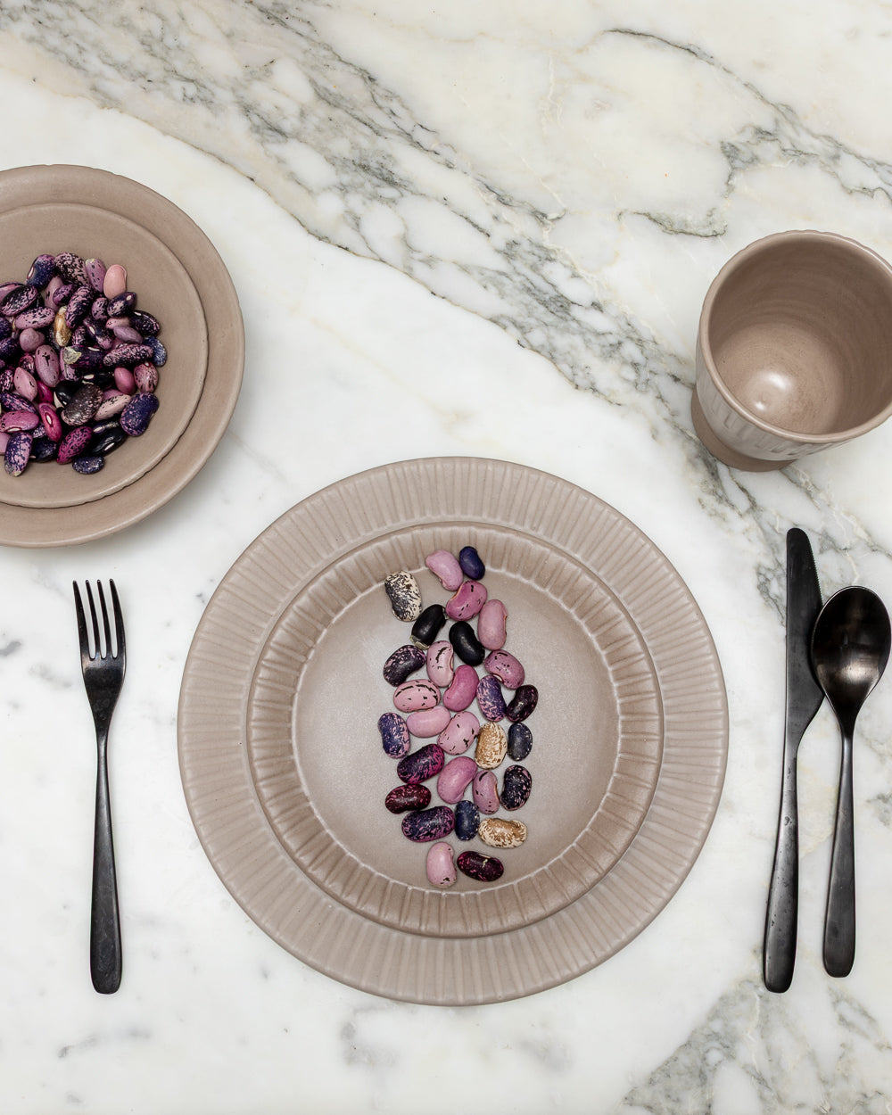 Place setting on white marble: clay-toned carved-rim plate with beans, black flatware and a matching cup