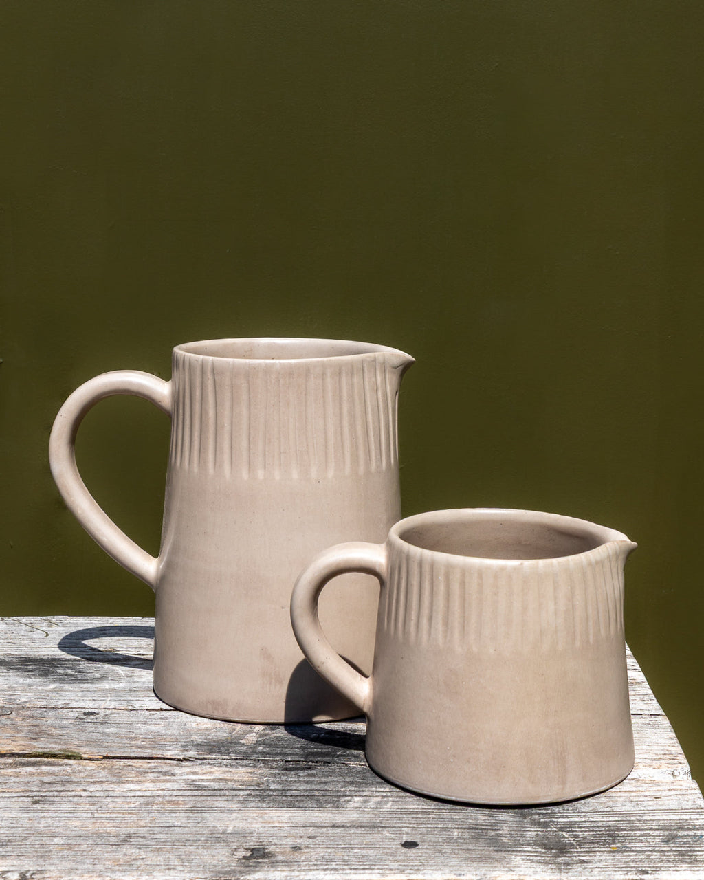Large and small clay-toned carved ceramic pitchers on a marble counter against a dark olive wall