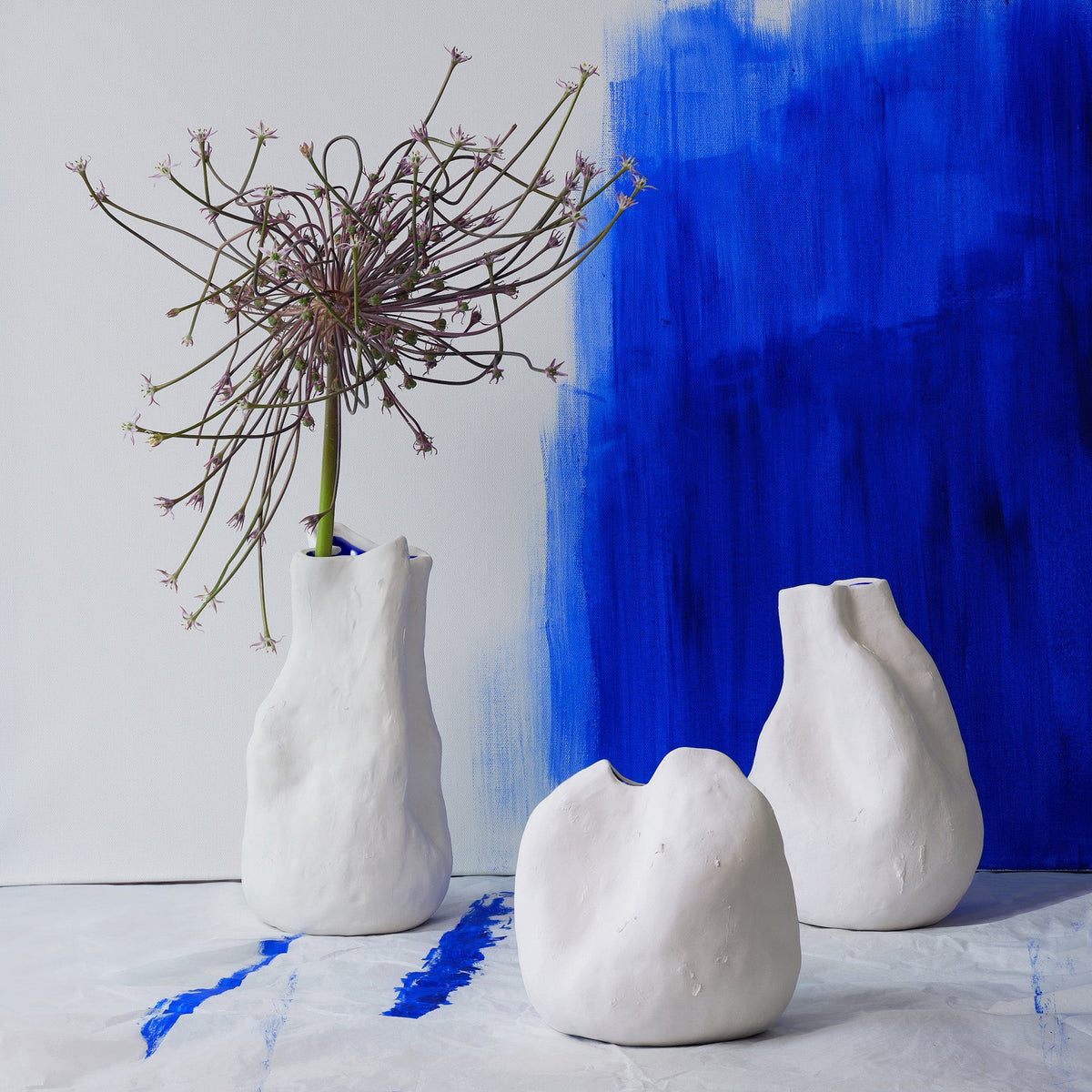 Three white sculpted porcelain vases, one holding a dried allium, in front of a cobalt brushstroke