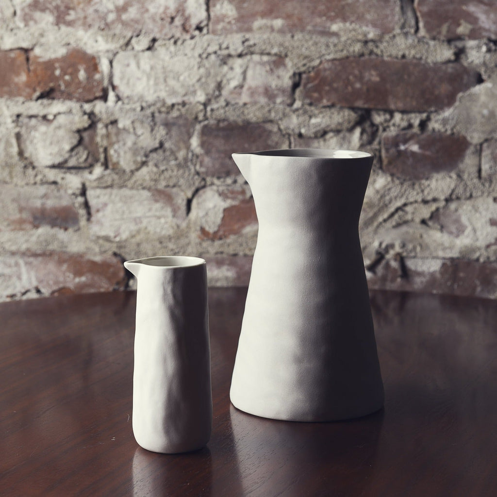 Tall pearl stoneware carafe beside a smaller pitcher on a dark wood table in front of an old brick wall