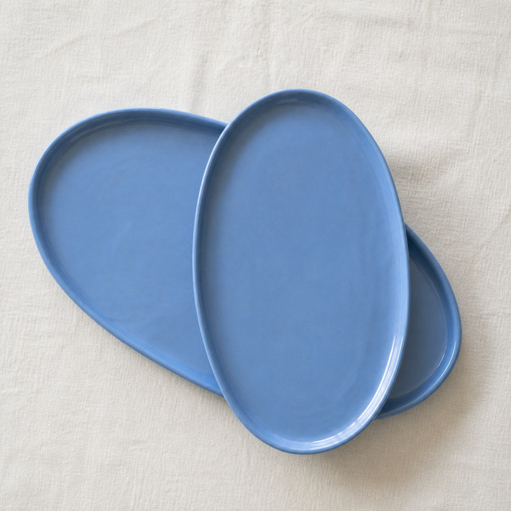 Three matte blue oval platters layered from large to small on a cream linen tablecloth