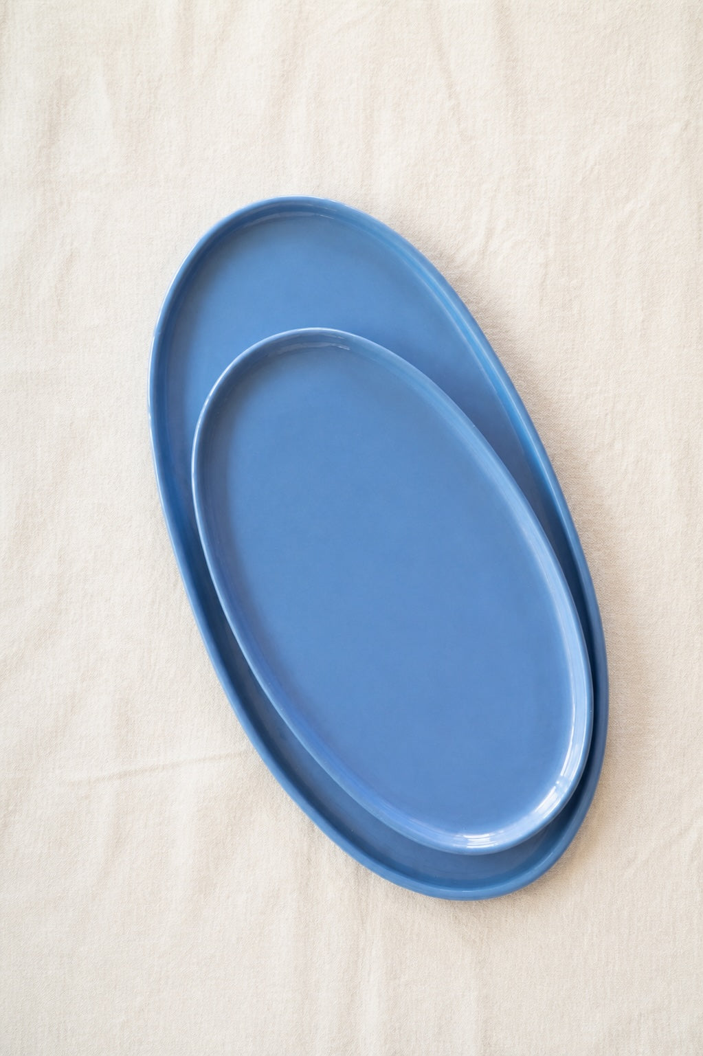 Large and medium blue porcelain oval platters stacked on white linen in soft daylight