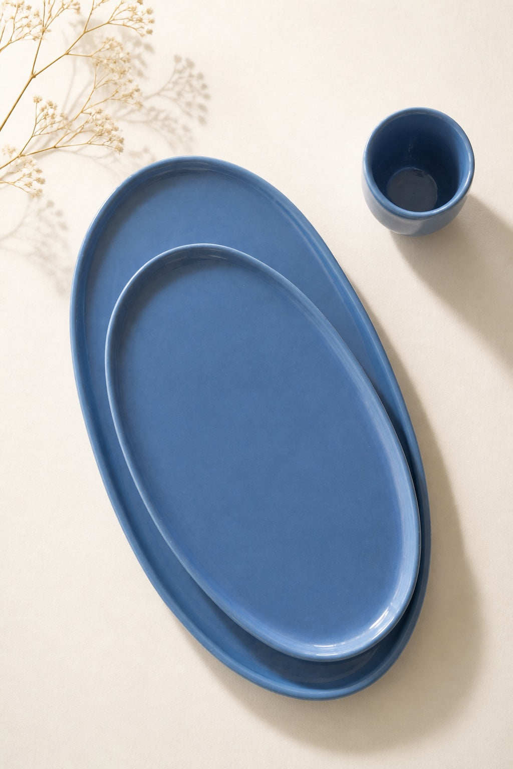 Nested blue oval porcelain platters beside a small blue cup and dried flowers on cream linen