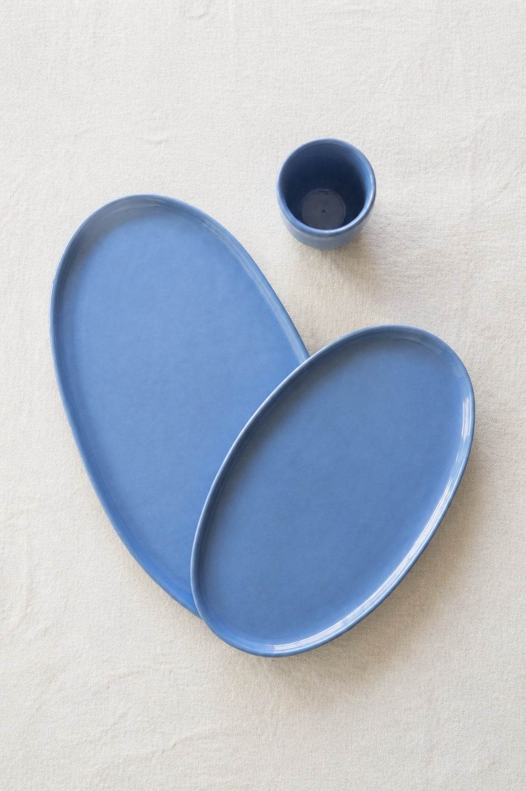 Two matte blue porcelain oval platters overlapping on white linen next to a small blue cup, seen from above