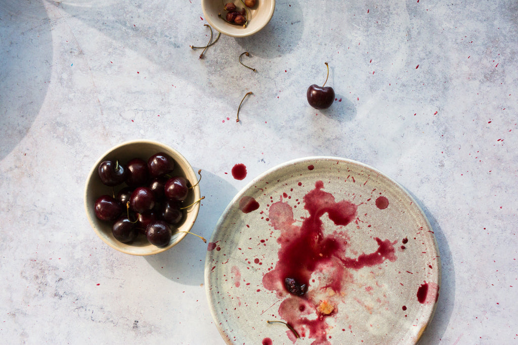 Horizon stoneware lunch plate in a pale oat glaze splashed with cherry juice beside a bowl of dark cherries