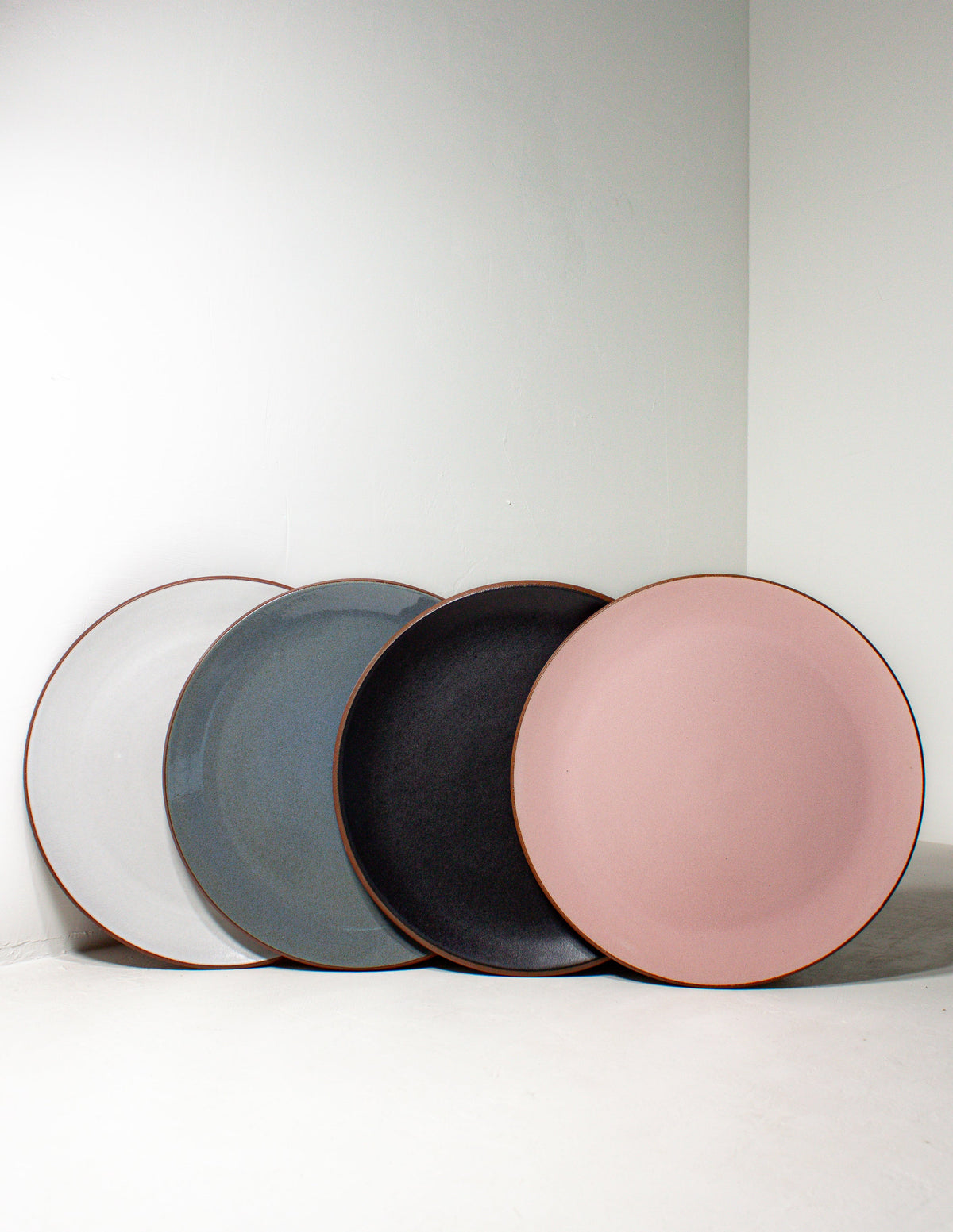 Four hand-thrown stoneware coupe dinner plates in white, sky blue, black and pink glaze leaning against a white wall