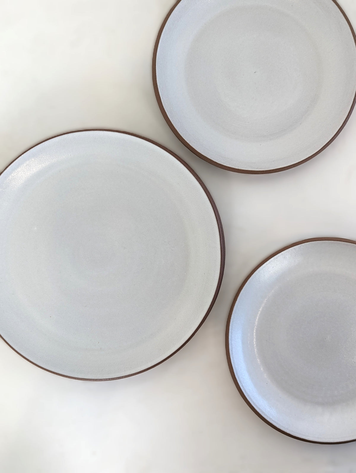 Three white glazed stoneware coupe dinner plates seen from above, bare clay rims overlapping on a pale surface