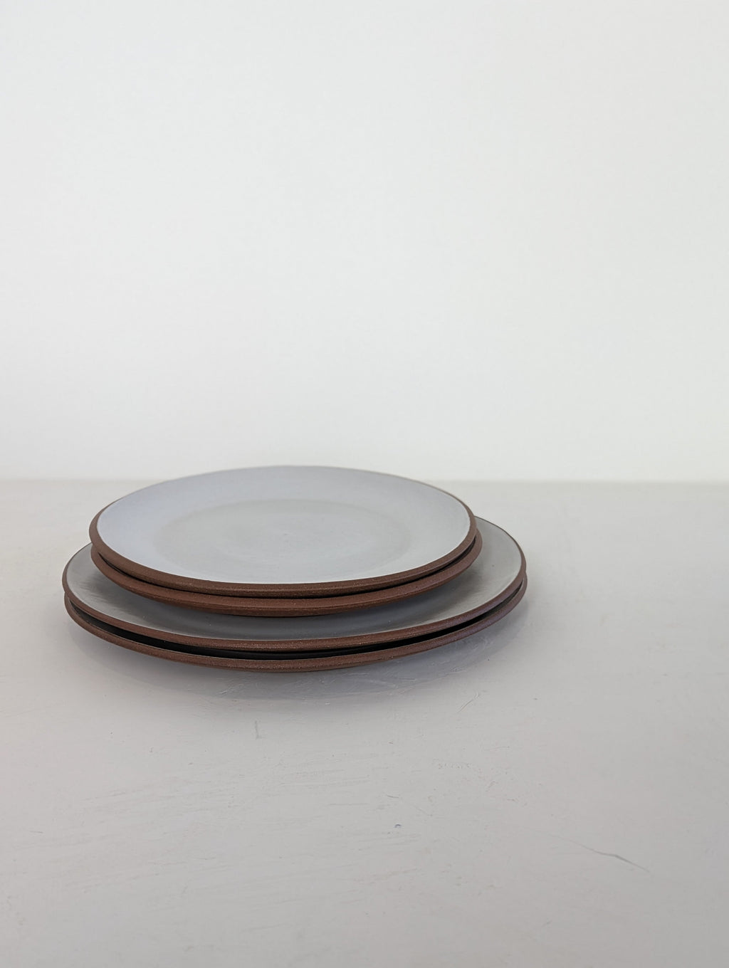 Two hand-thrown stoneware coupe plates in pale grey white glaze stacked on a white surface, dinner size on the bottom