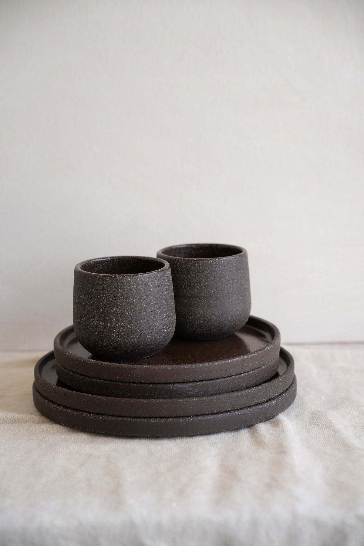 Pair of stoneware coffee cups in speckled dark brown set on four stacked matching plates on a linen tablecloth
