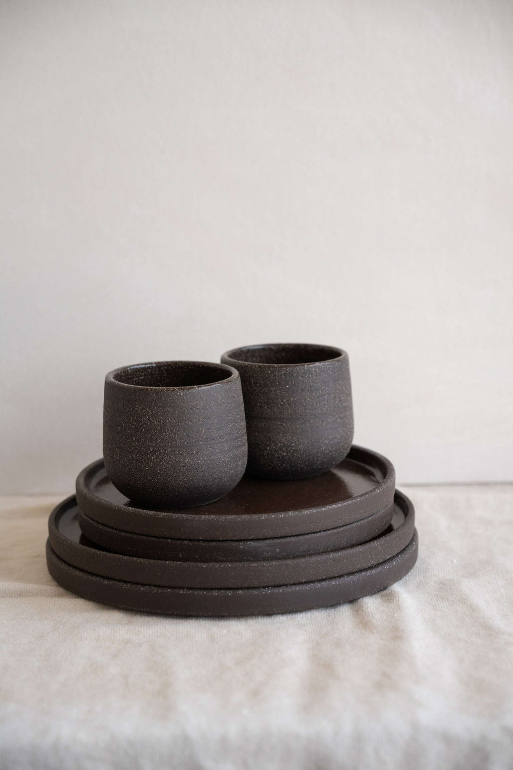 Stoneware coffee cups in speckled dark brown, two side by side on a short plate stack against a pale plaster wall