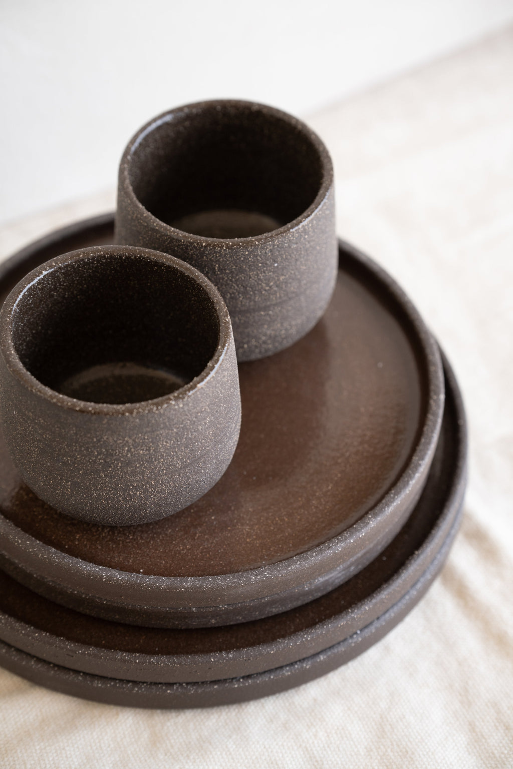 Two stoneware coffee cups in speckled dark brown on a stack of matching plates, seen from above at an angle