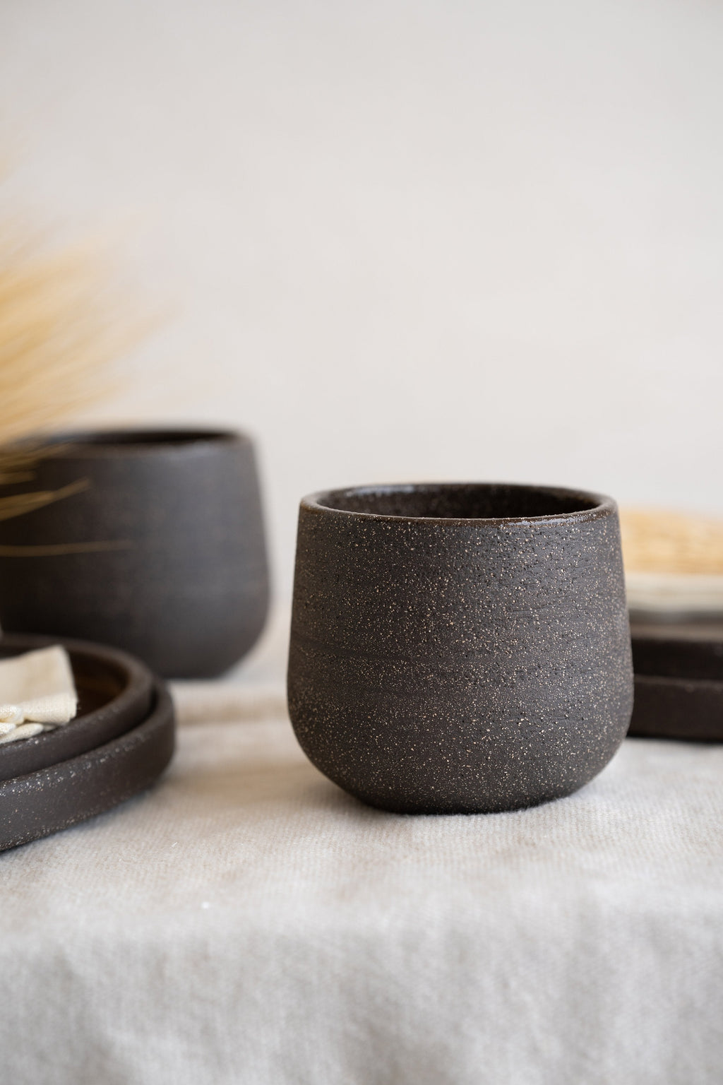 Stoneware coffee cup in speckled dark brown, close side view on grey linen with matching cup and plates blurred behind