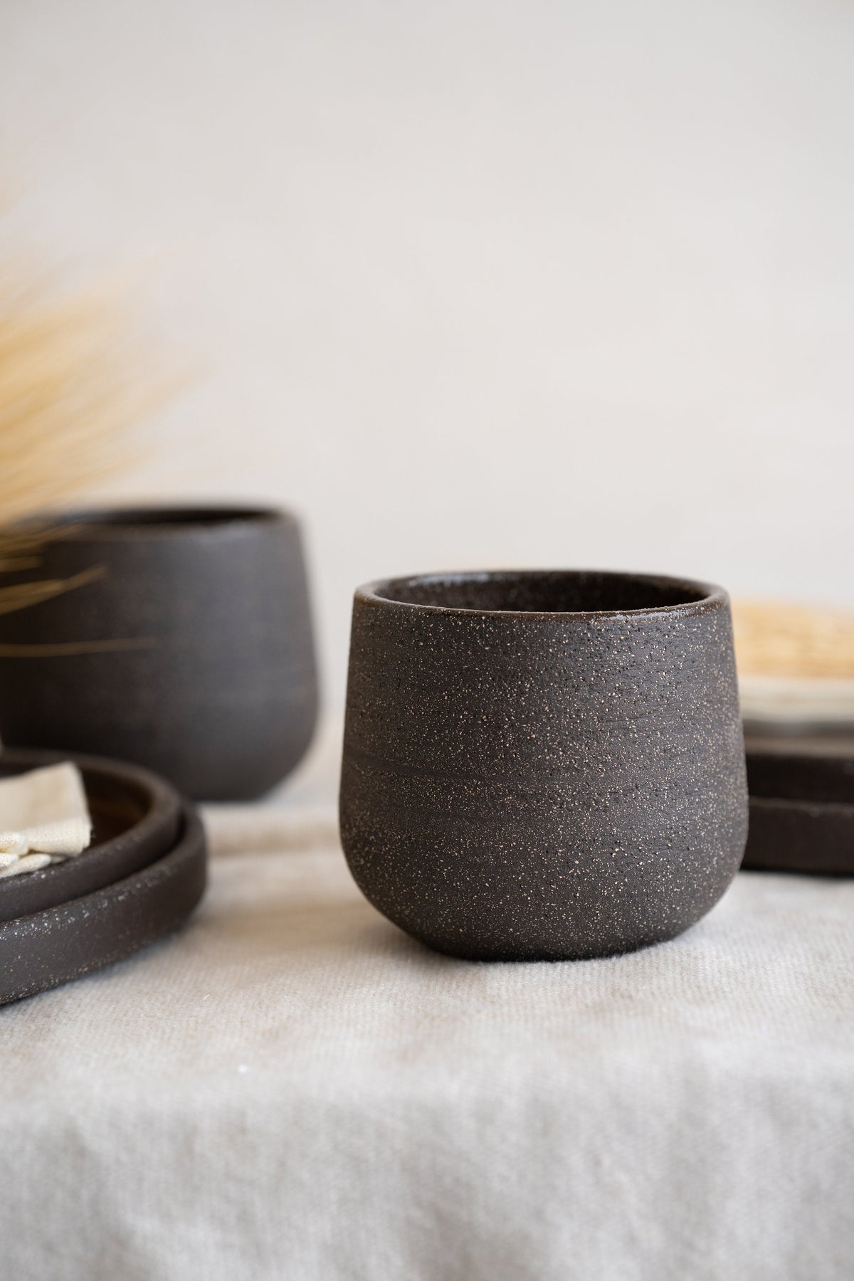 Stoneware coffee cup in speckled dark brown, close side view on grey linen with matching cup and plates blurred behind