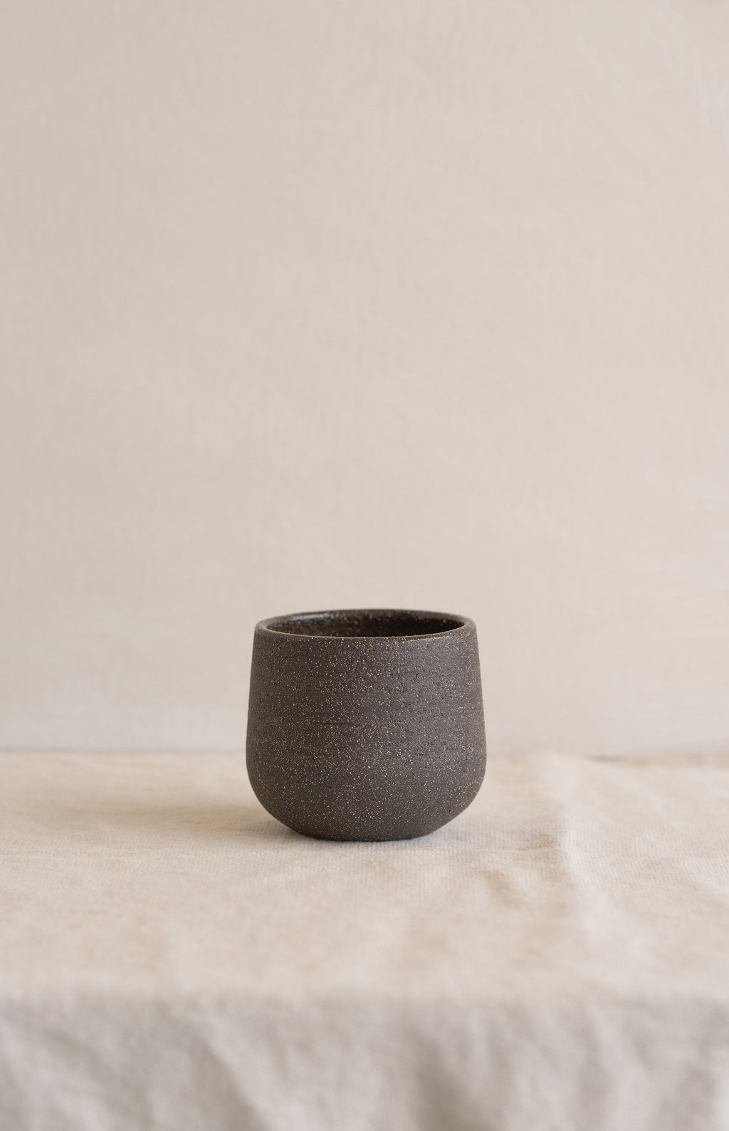 Handleless stoneware coffee cup in speckled dark brown matte clay standing alone on a cream linen cloth, front view