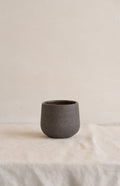 Handleless stoneware coffee cup in speckled dark brown matte clay standing alone on a cream linen cloth, front view