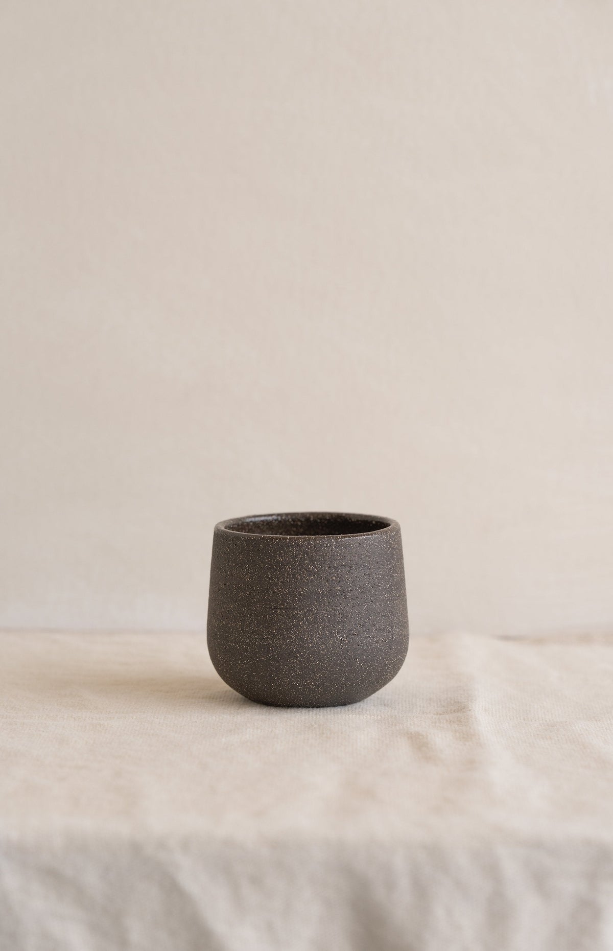 Handleless stoneware coffee cup in speckled dark brown matte clay standing alone on a cream linen cloth, front view