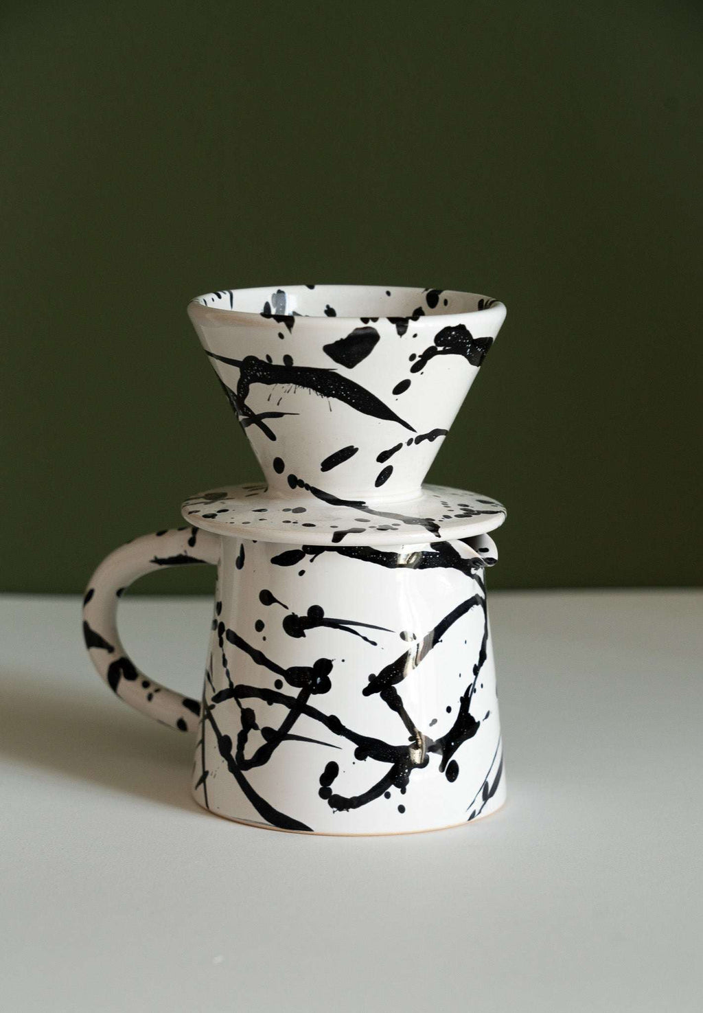 Splatter glazed stoneware pour-over dripper stacked on a matching pitcher against a dark olive green wall