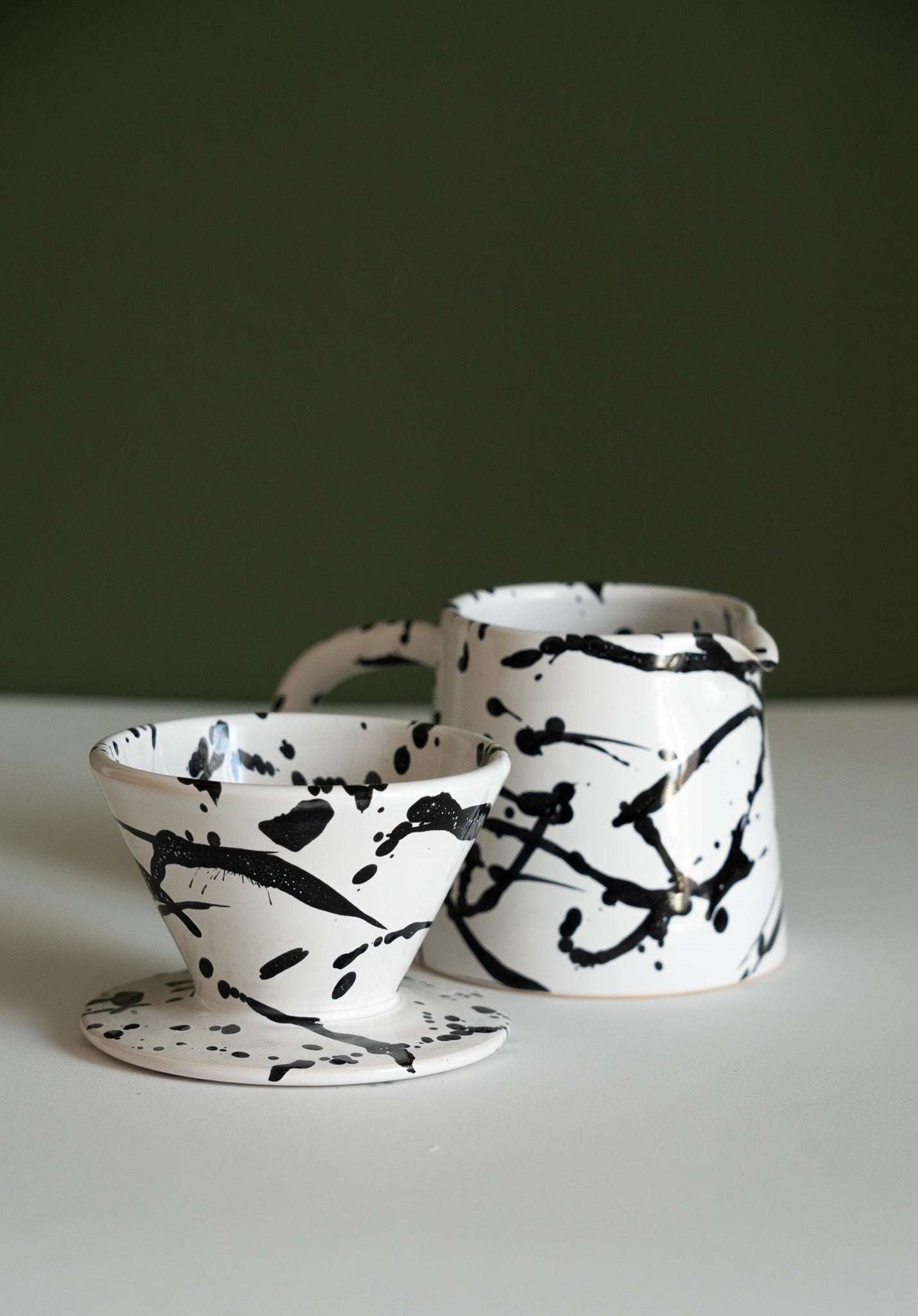 Splatter stoneware pitcher behind its cone dripper lying on its side on a white tabletop
