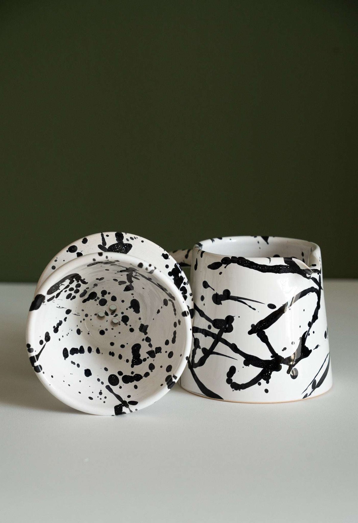 Speckled stoneware dripper on its side beside the pitcher on a white surface and green wall