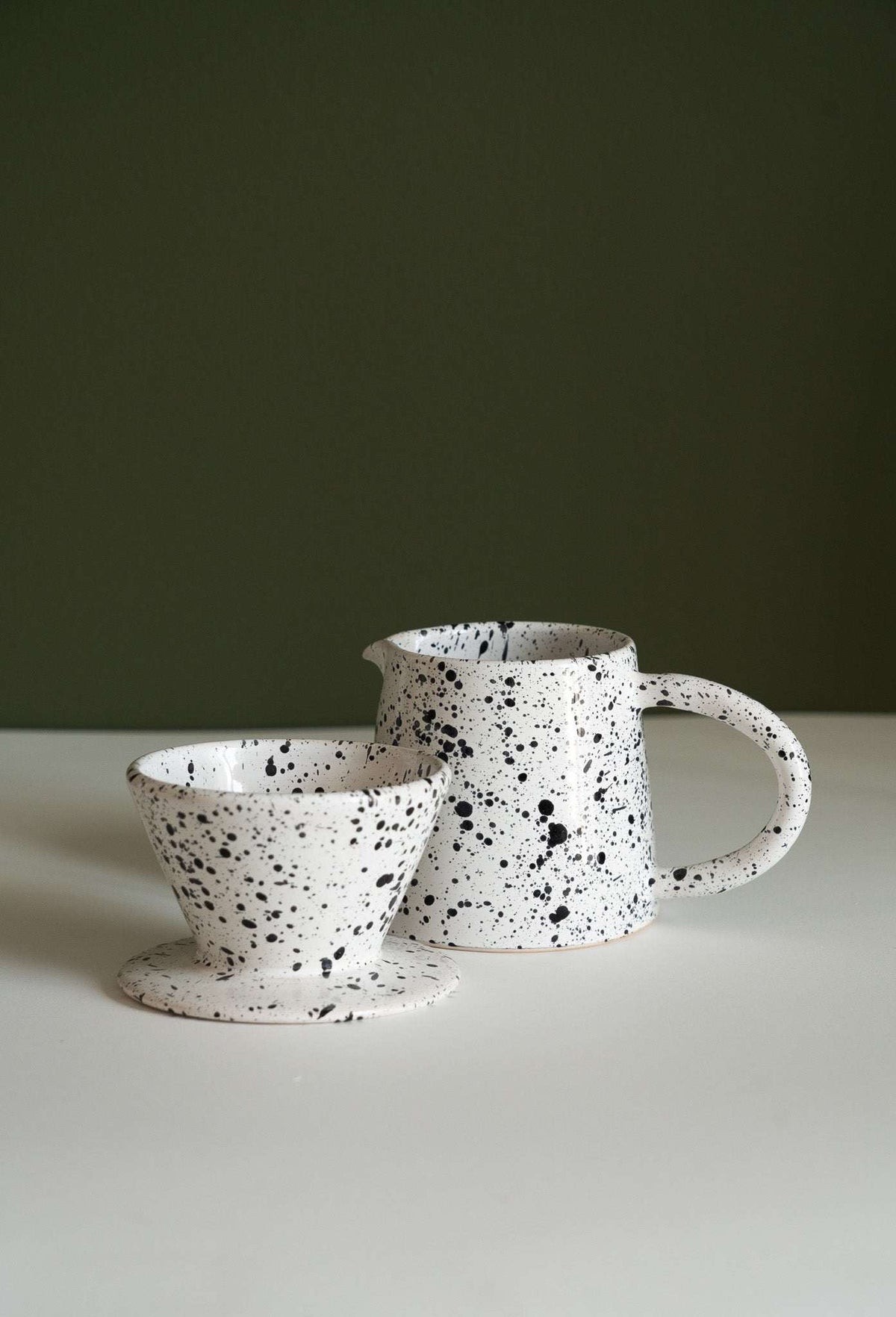 Fine speckle stoneware dripper and pitcher side by side on a white tabletop and green wall