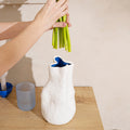 Hands lowering a bunch of green stems into a tall white sculpted porcelain vase on a wood counter