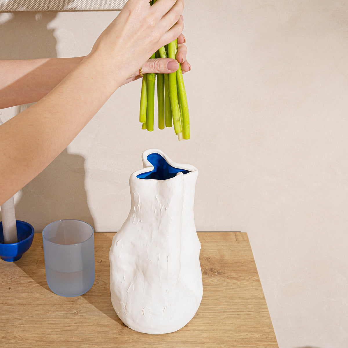 Hands lowering a bunch of green stems into a tall white sculpted porcelain vase on a wood counter