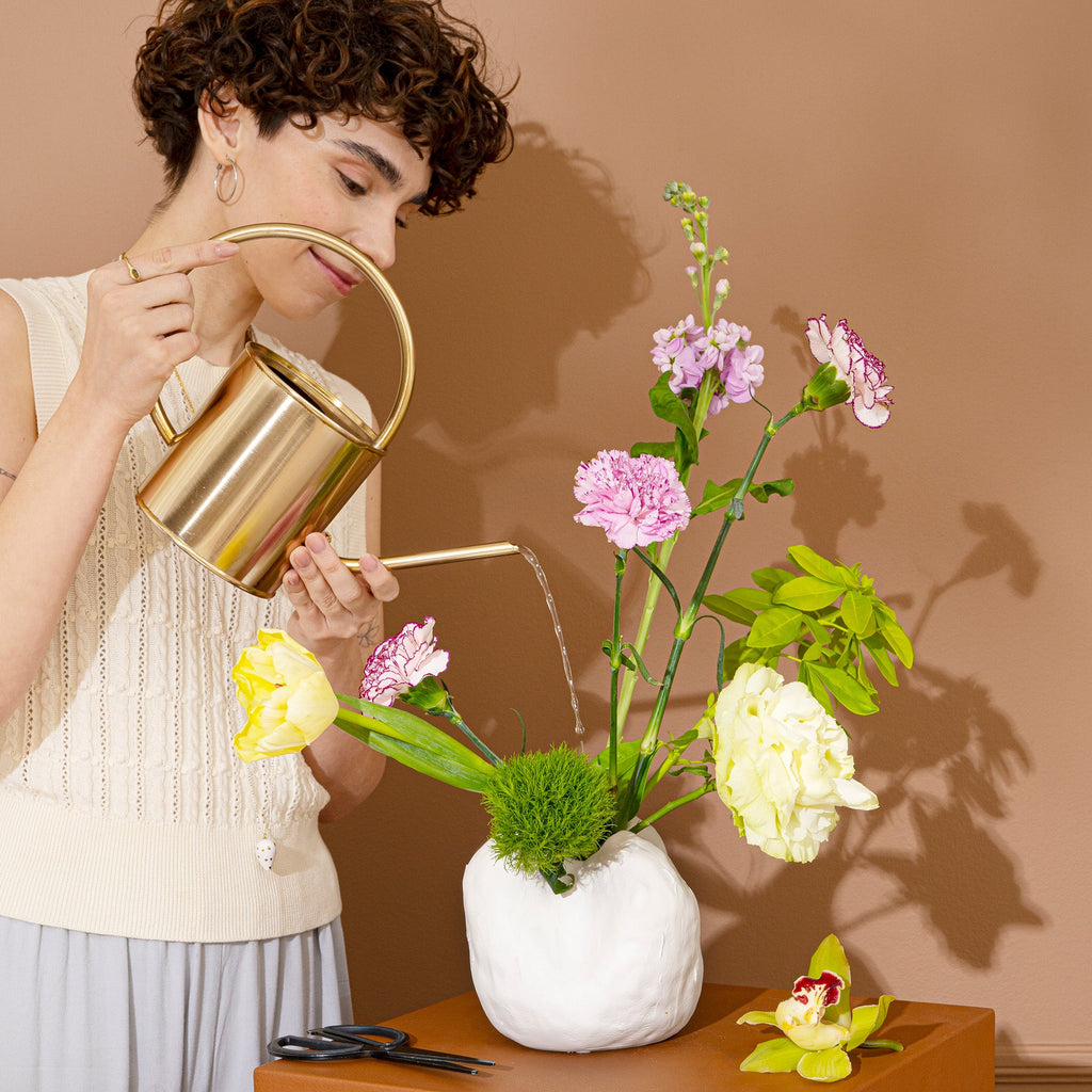 Woman watering pink and green flowers arranged in a small white sculpted porcelain vase on a wood table