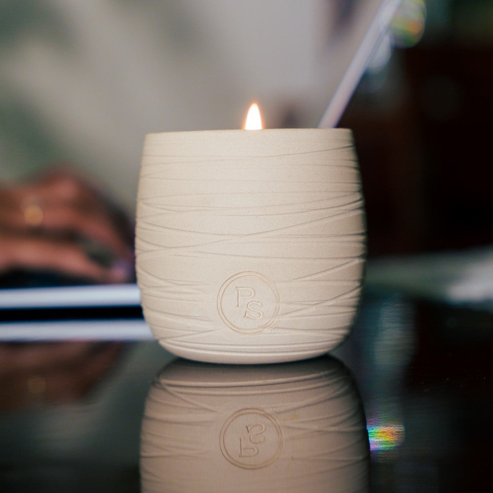 Lit neroli candle in a carved cream clay vessel on a dark glass table with a laptop softly blurred behind