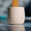Neroli and basil candle in a ridged sand-colored clay vessel with an embossed seal, reflected on a glossy table
