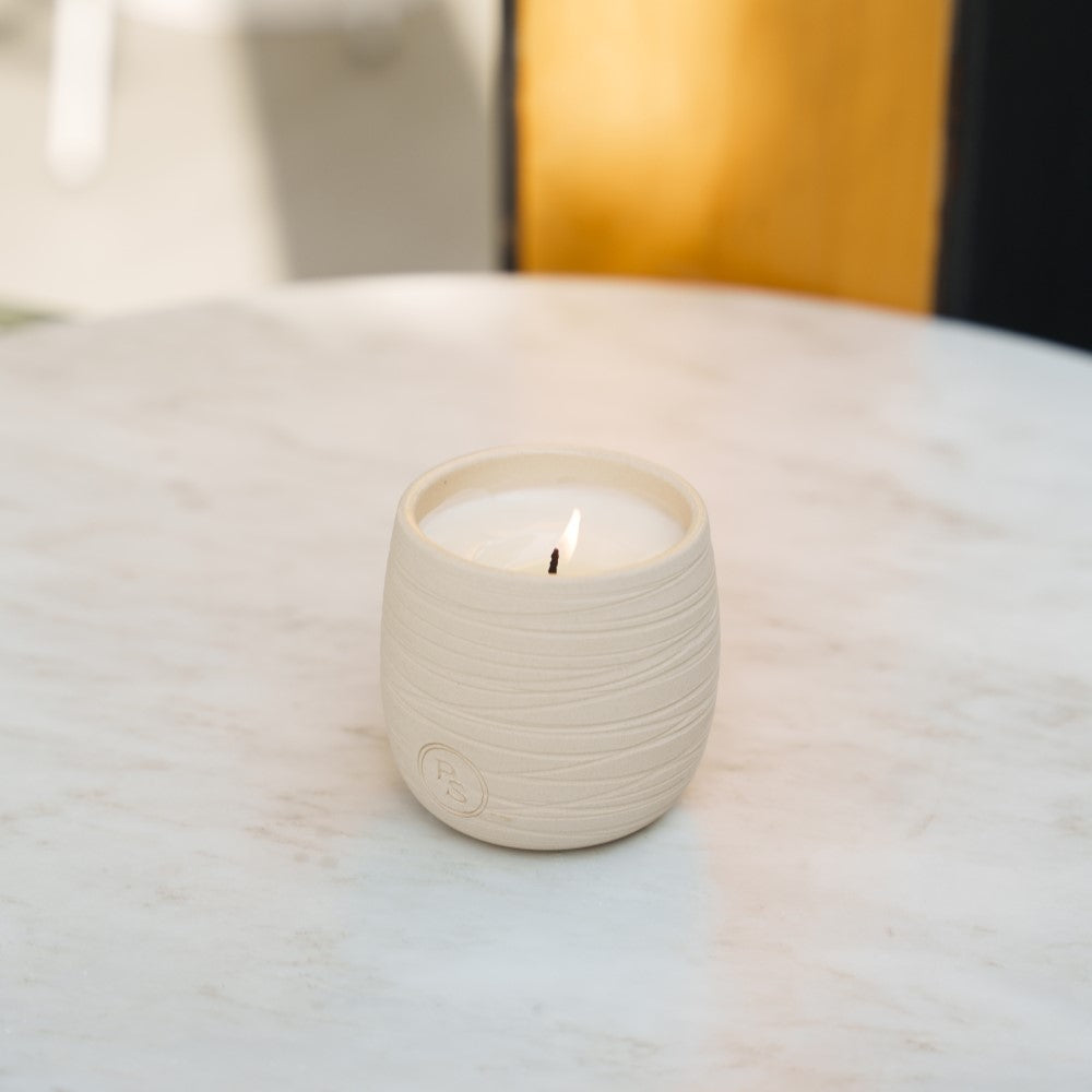 Small lit neroli candle in a pale handmade clay vessel on a white marble table with warm light behind