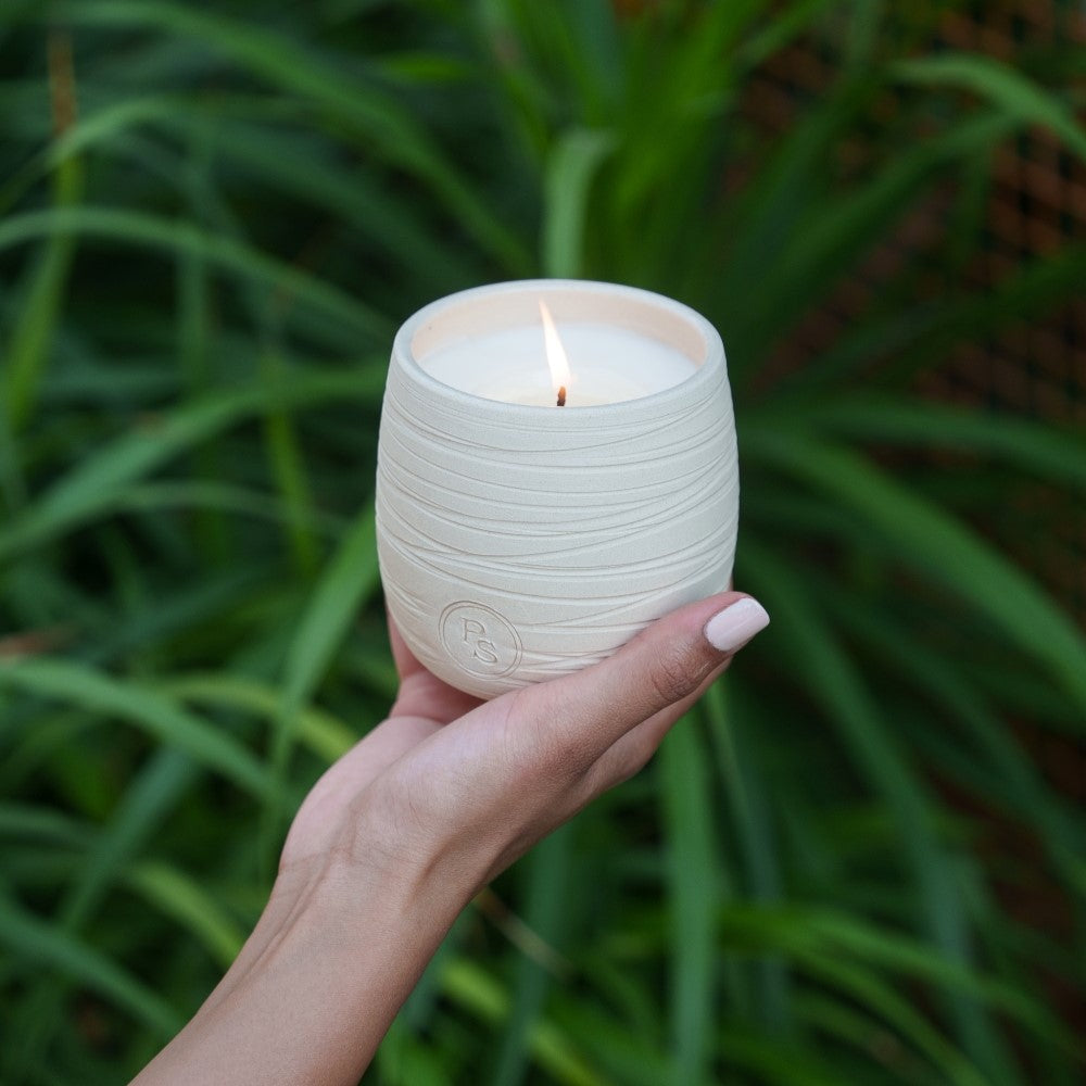 Hand holding the lit neroli soy candle in its ridged white clay vessel in front of long green leaves